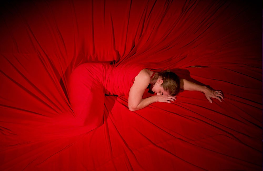 A woman lies on a floor with a red cloth draped over her body and floor. 