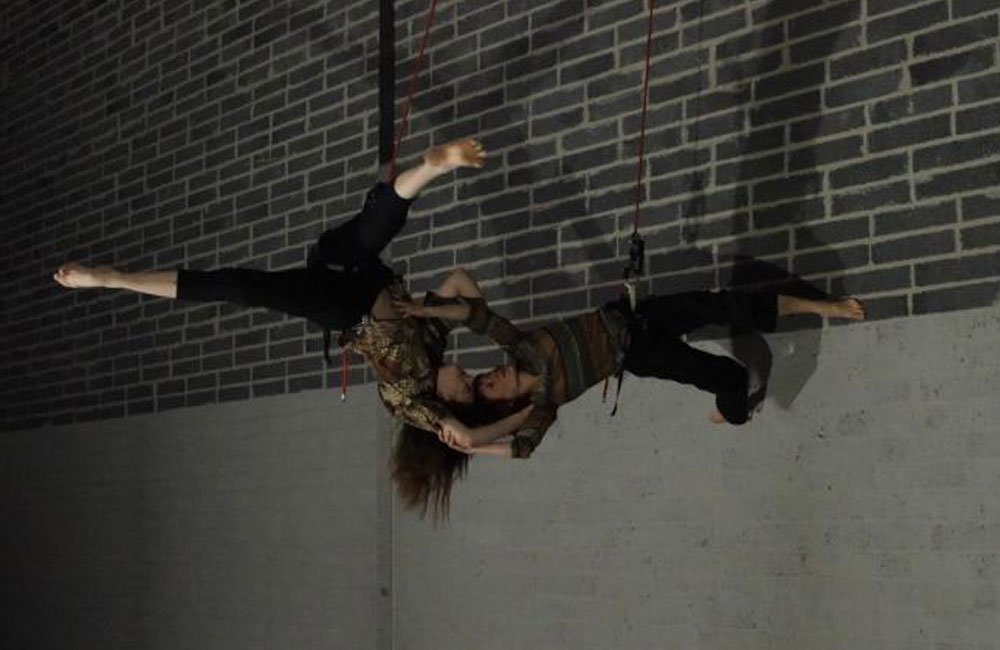 Two aerialists perform against a concrete and brick wall.