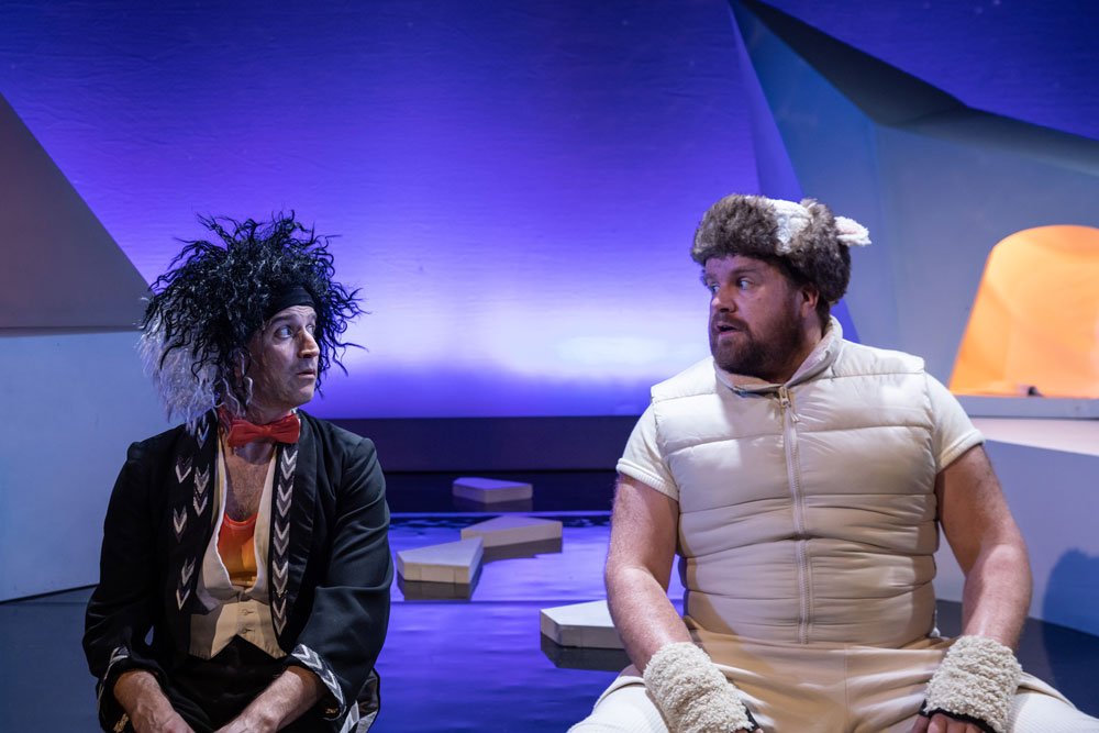A photo of two actors looking at each on a theatre stage.  The man on the left has wild black hair and is wearing a scruffy back jacket with arrows drawn on. The man on the right is all in white and is wearing a fur and and gloves, a short sleeved tee-shirt and a padded jacket.