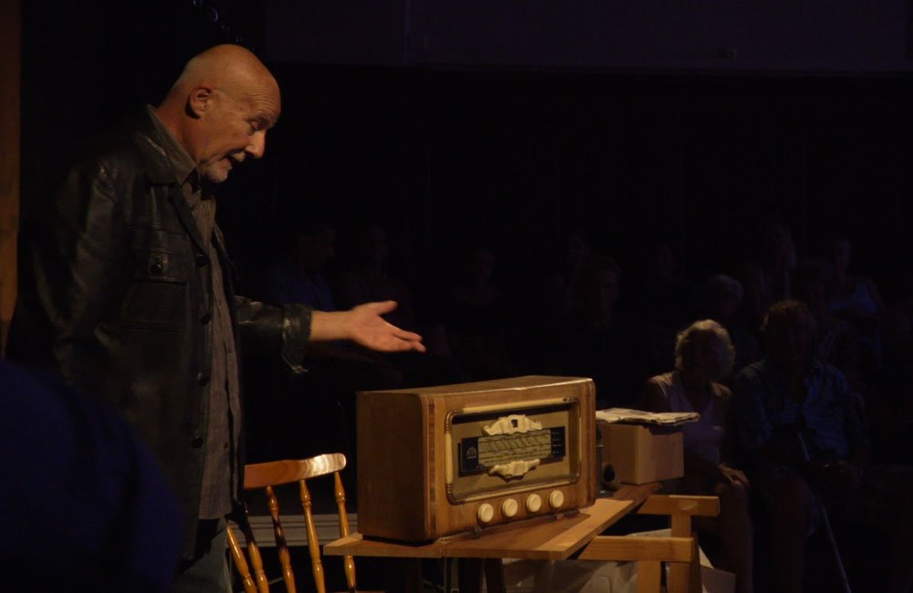 An older man gestures towards a vintage radio sitting on a rustic table on a darkly lit theatre stage.