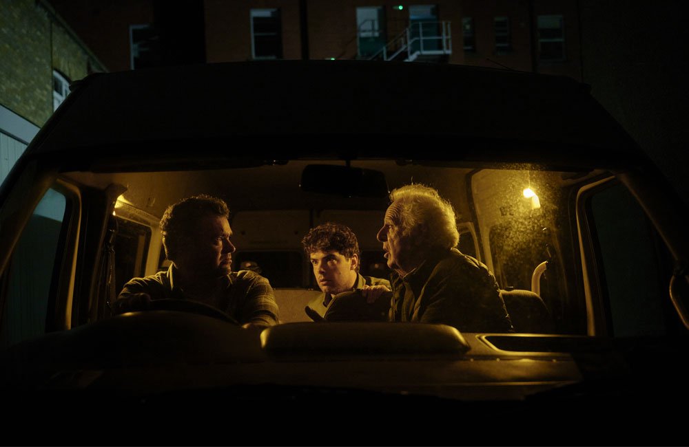 Three men engaged in a tense conversation in the dim, night time interior of a minibus.
