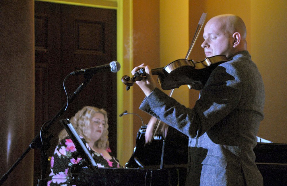 A male plays the fiddle to the accompaniment of a female pianist.