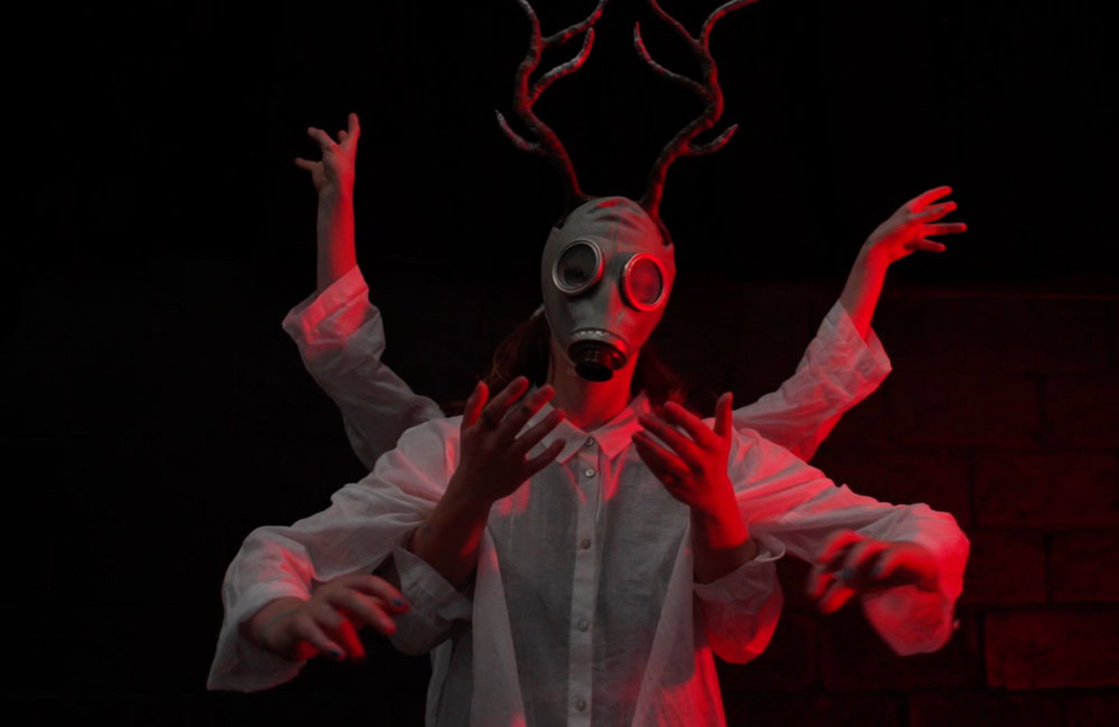 A performer wearing a gas mask under dramatic red lighting. They are wearing deer antlers on their head. Two other performers are using their arms to create the impression that the main perforrmer in multi-limbed.