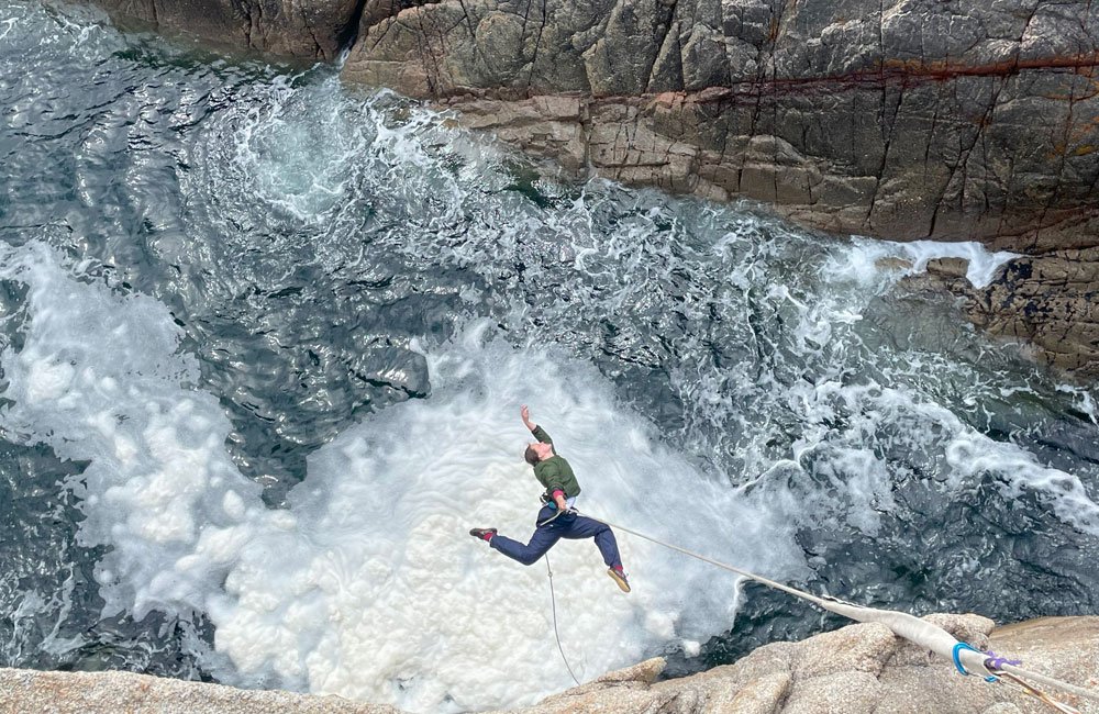 An aerial dancer performs on a cliff face over crashing waves.