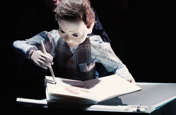 A photo of a hand crafted theatre puppet. He is wearing a shirt and a tweed waistcoat and is drawing something in a book.