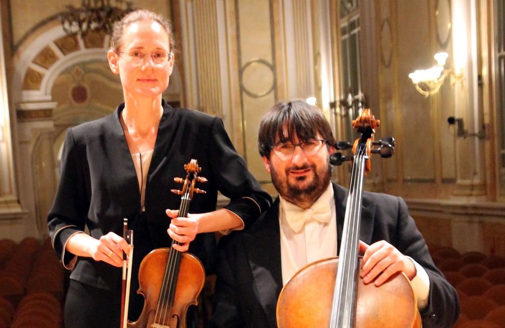 A woman holding a violin stands beside a seated man with a cello. They are both white and wearing formal evening wear.