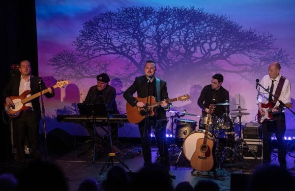 A five piece band perform on a theatre stage. The back drop is a projection of winter trees against a blue and purple evening sky.