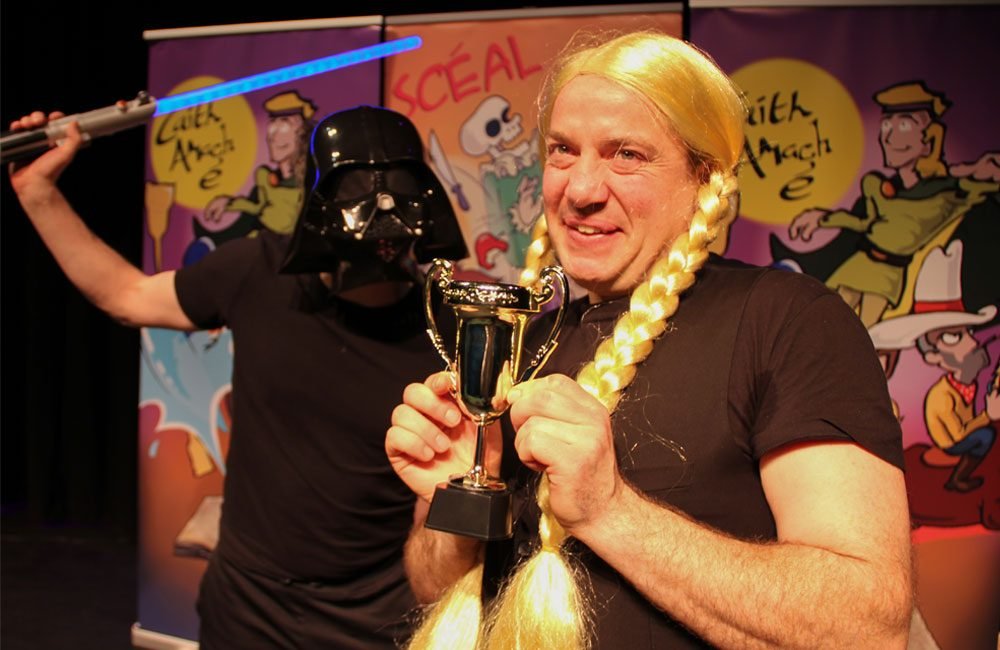A man wearing a blonde wig with long plaits holds a tiny trophy. Behind him a man wearing a Darth Vader helmet brandishes a toy lightsaber.