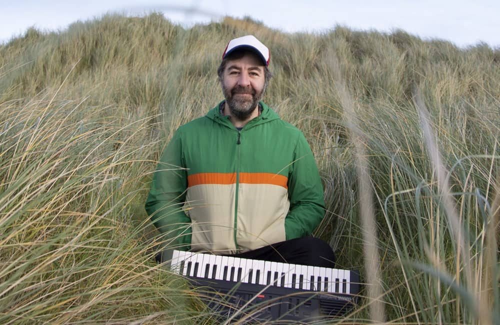 Comedian David O'Doherty standing in tall grass. He is wearing a baseball hat and a green and cream hoodie with an orange stripe. An electronic piano keyboard is in front of him.