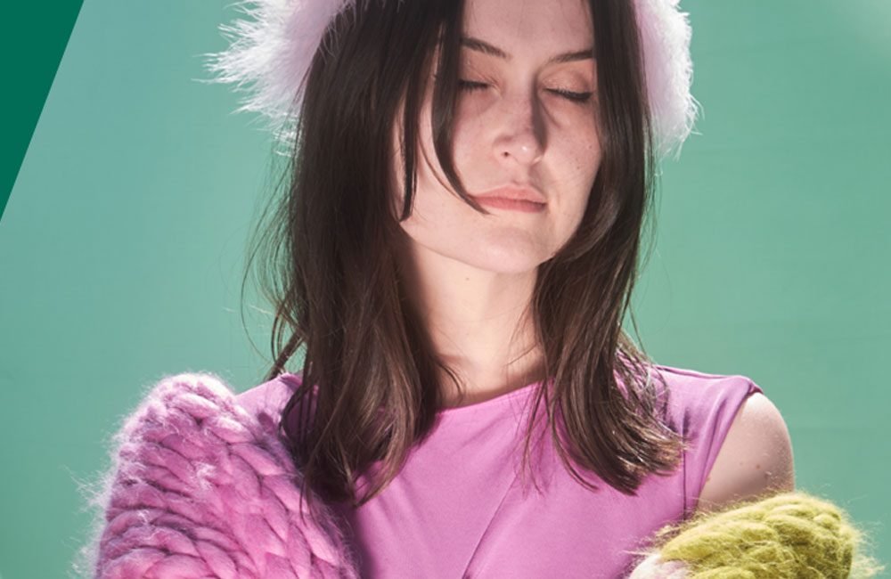 A young white woman with long dark hair. Her eyes are closed. she is wearing pink clothes against a green background.
