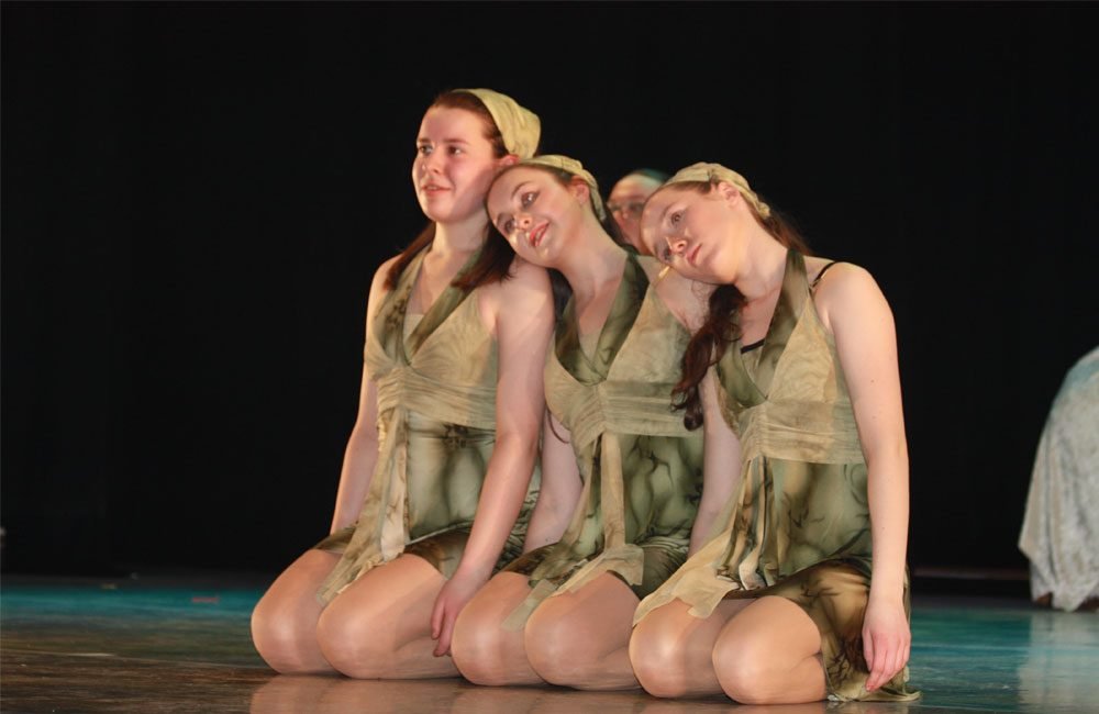 4 young dancers kneel together on a stage, They are all wearing matching green costumes and headbands.