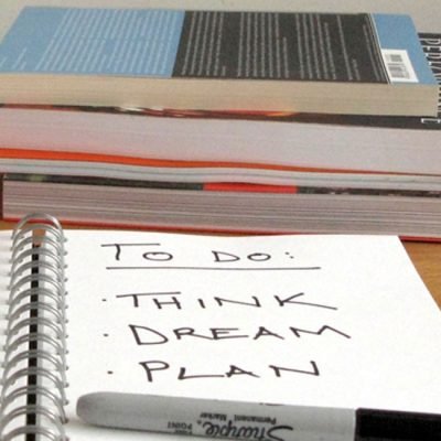 An open notebook sits on a desk in front of a stack of books. The words: To do: think, dream, plan are written in the notebook with a sharpie pen.
