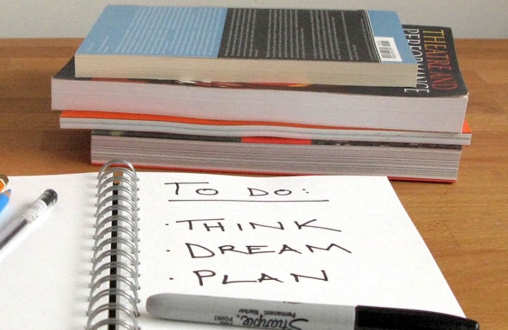 An open notebook sits on a desk in front of a stack of books. The words: To do: think, dream, plan are written in the notebook with a sharpie pen.