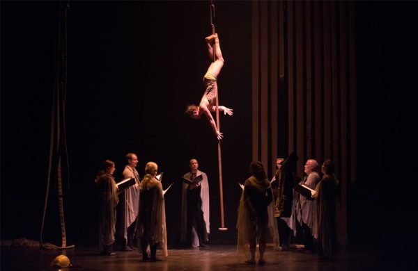 An aerial performer hangs in an inverted pose above a group of performers holding open books as if they are reading or singing.