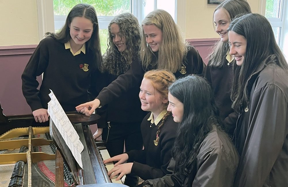 A group of teenage girls wearing brown school uniforms gather around a piano.