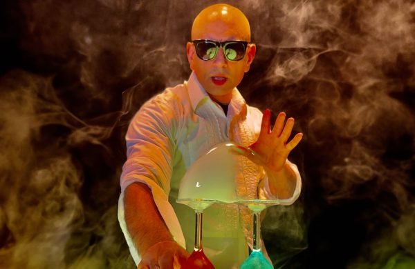 Photo of a bald white man wearing sun glasses and a lab coat. He is surrounded by swirling mist. In front of him are some up turned wine glasses covered in a large soap bubble.