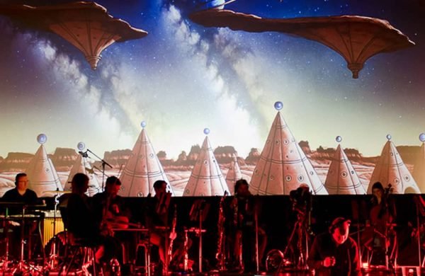 An orchestra performs in from of a surreal projection of wigwam tents on a theatre stage.
