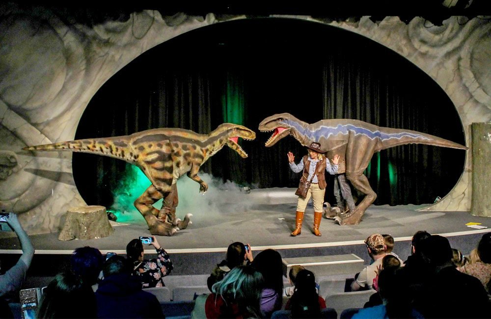 A woman dressed in safari gear performs on a theatre stage with two large dinosaurs.
