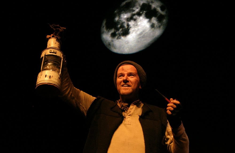 A man dressed as a 19th century sailor holds a storm lantern up with one hand. The full moon is visible behind him.